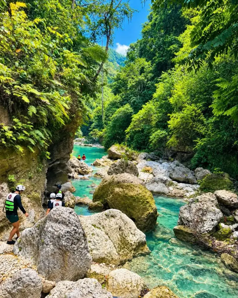 Canyoneers in helmets climbing over pale boulders beside a turquoise river in a jungle canyon