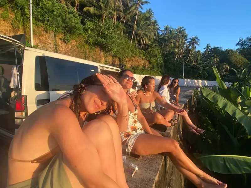 Guests sitting on a roadside wall in golden-hour light beside the tour van and coconut palms