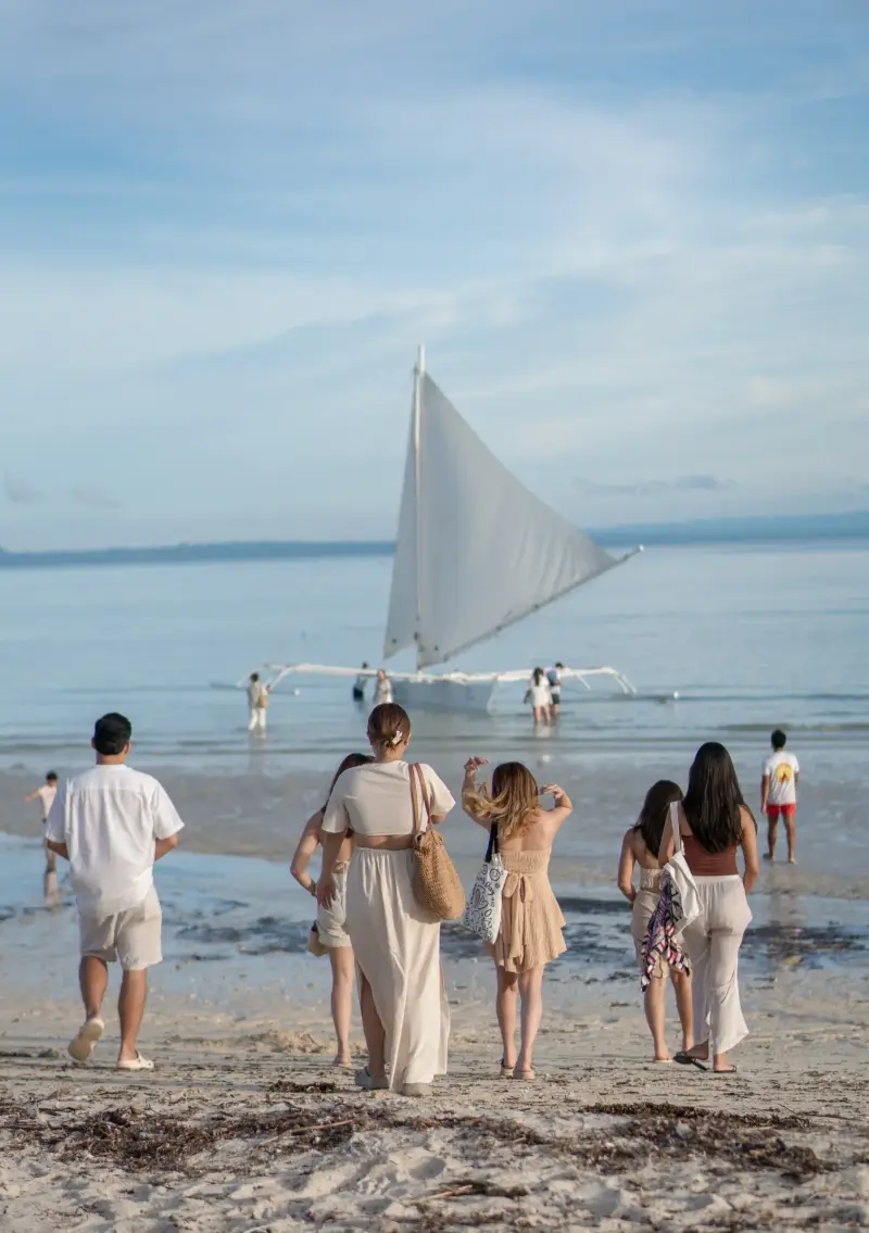 Guests walking across the sand towards a paraw with its white sail raised in the shallows
