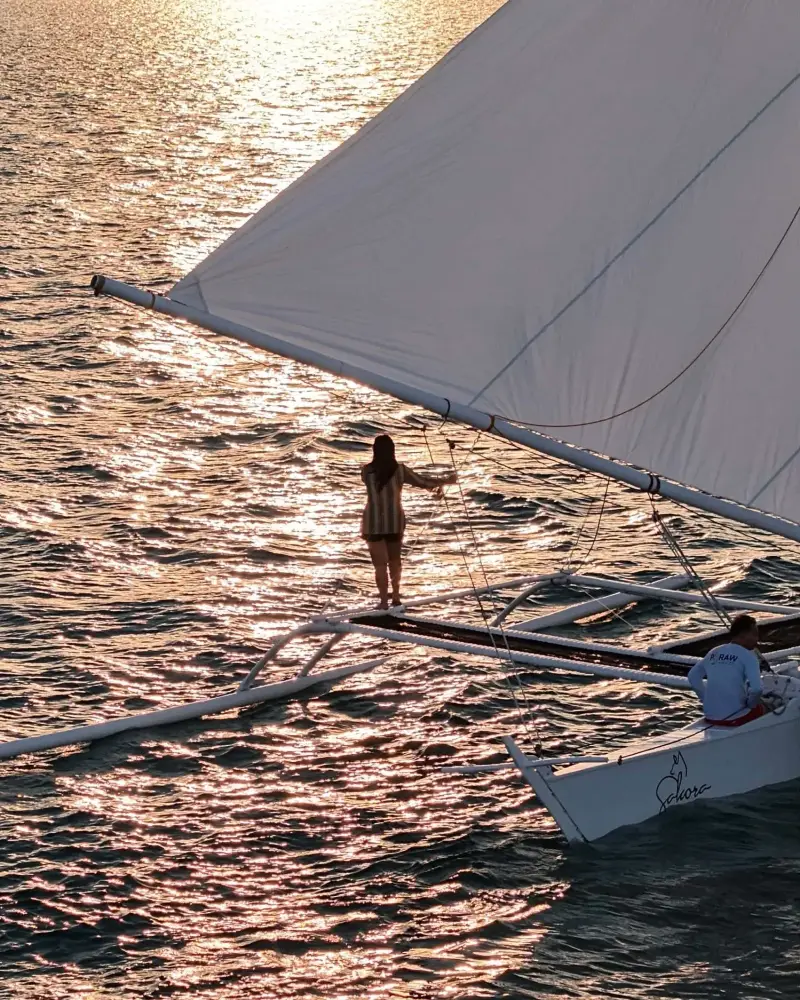 A guest standing on the outrigger beside a sail lit gold by the late-afternoon sun