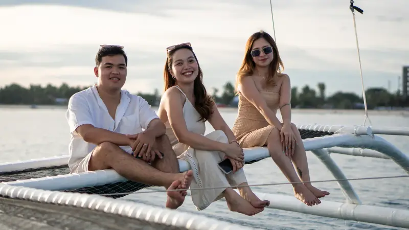 Three guests sitting on the paraw's bamboo outrigger, smiling, with the shore behind them
