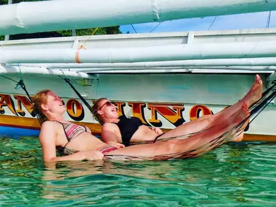 Two guests floating in clear water beside a traditional paraw sailboat