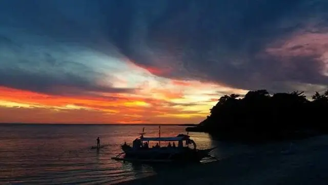 A bangka anchored off Malapascua's wooded shore under a red and orange sunset